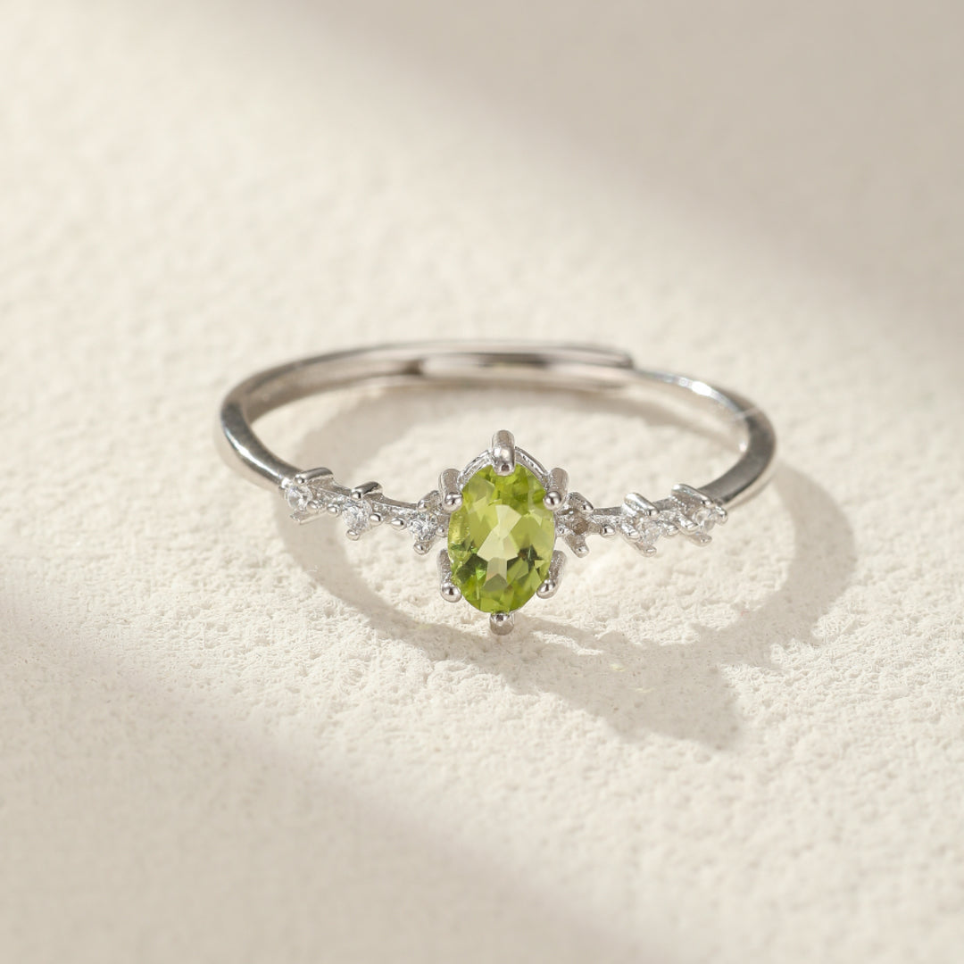 Bague en argent sertie d'une pierre de péridot ovale verte, avec de petites pierres blanches incrustées sur les côtés, sur un fond texturé clair.