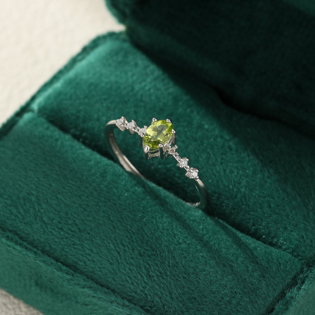 Bague en argent ornée d'une pierre centrale ovale péridot vert clair, entourée de petites pierres blanches scintillantes, posée sur un écrin en velours vert.