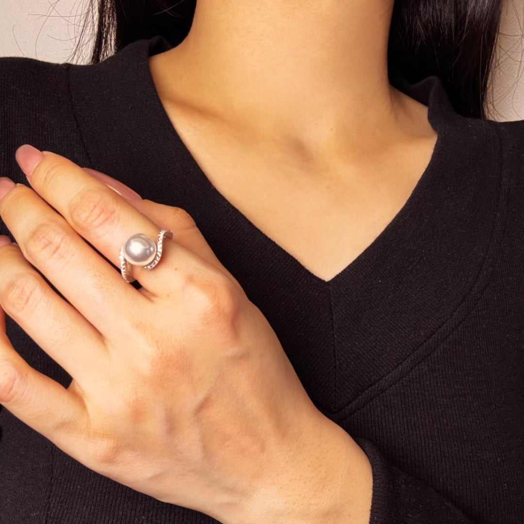 Bague en argent sertie d'une perle grise centrale et de petits cristaux scintillants sur un anneau torsadé, portée au doigt par une femme.