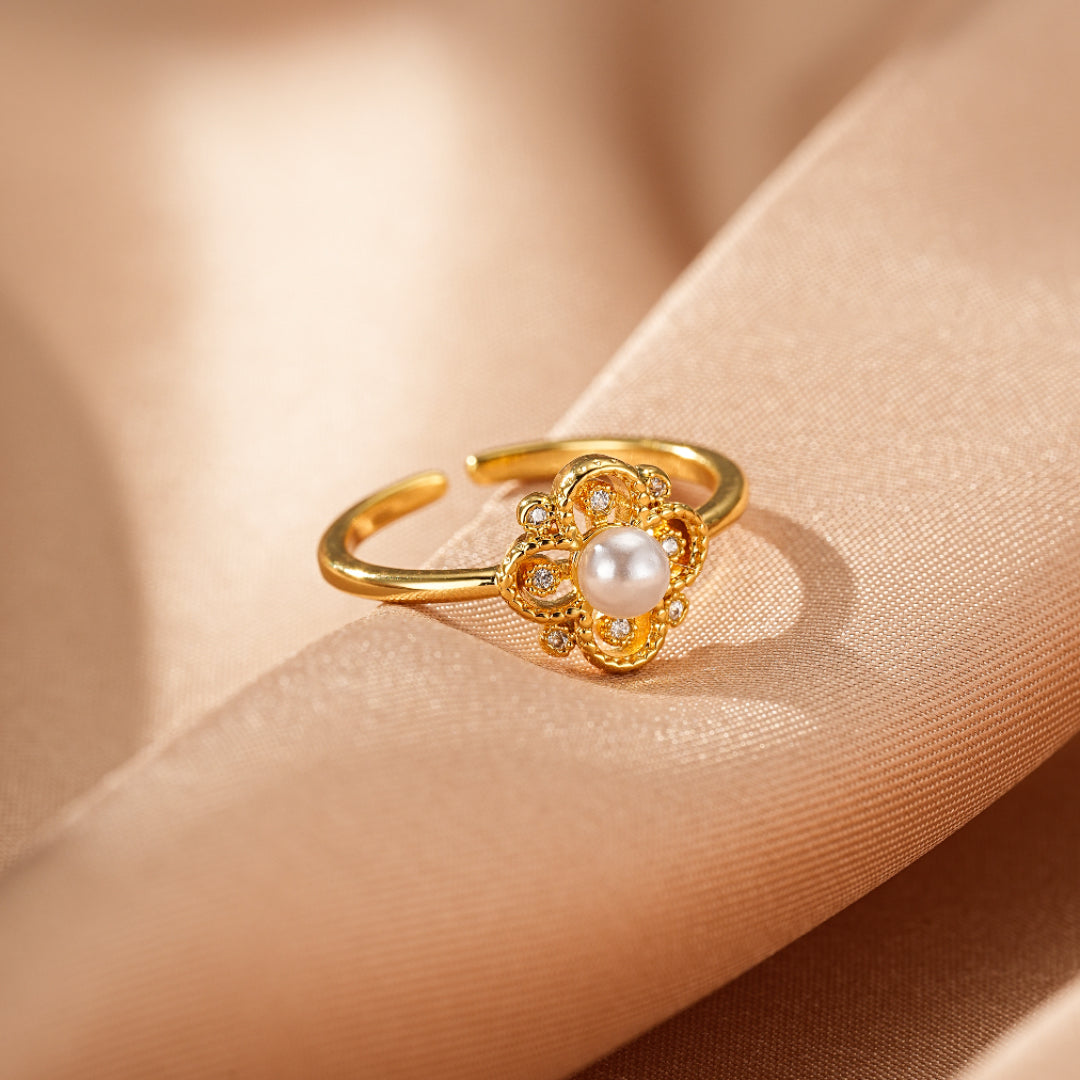 Bague en or ajustable ornée d'une perle nacrée au centre et de petits cristaux scintillants disposés en forme de fleur de trèfle sur un fond satiné beige.
