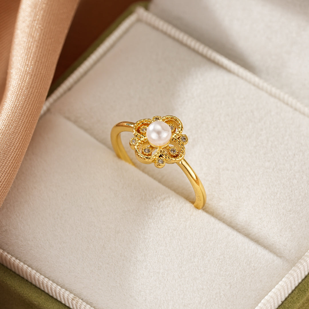 Bague en or jaune avec une perle blanche au centre, entourée de petites pierres scintillantes formant une fleur stylisée. La bague est présentée dans un écrin écrin vert.
