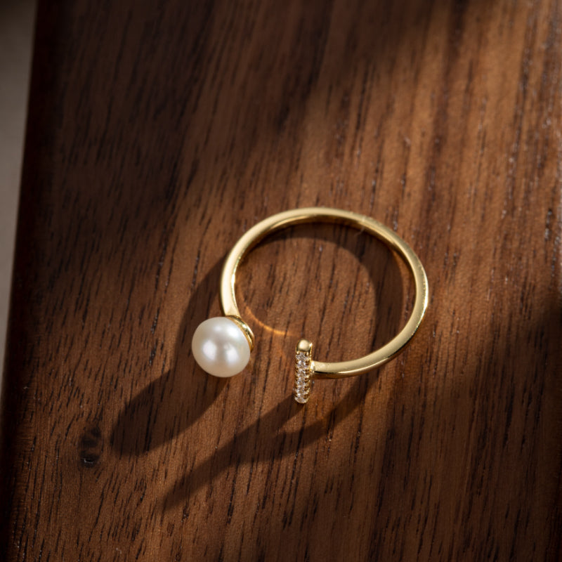 Bague ouverte plaquée or avec une perle blanche et une barre de zircons cubiques, posée sur un fond en bois sombre.