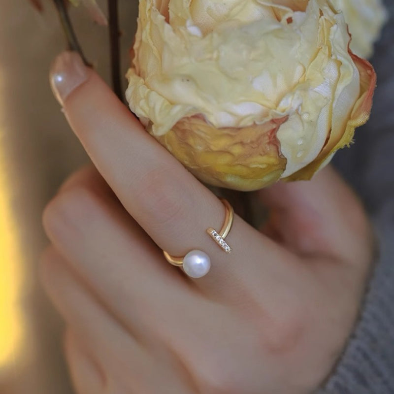 Bague ouverte en plaqué or avec une perle et de petits diamants incrustés, portée à la main près d'une fleur séchée.