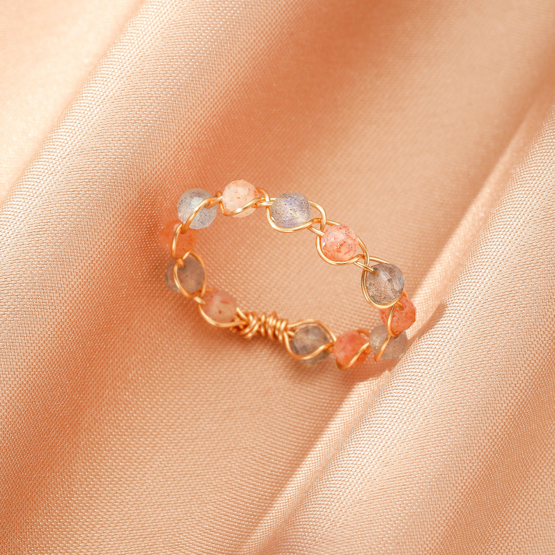 Bague Éclat Céleste artisanale en fil doré avec perles de pierre de soleil orange et pierre de lune aux reflets gris, présentée sur un fond en satin beige.