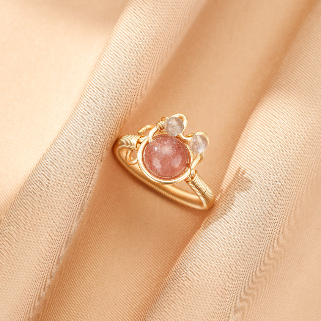 Bague en filigrane doré sertie d'une pierre rose ronde et de deux petites pierres claires, sur un fond de tissu satiné couleur pêche.