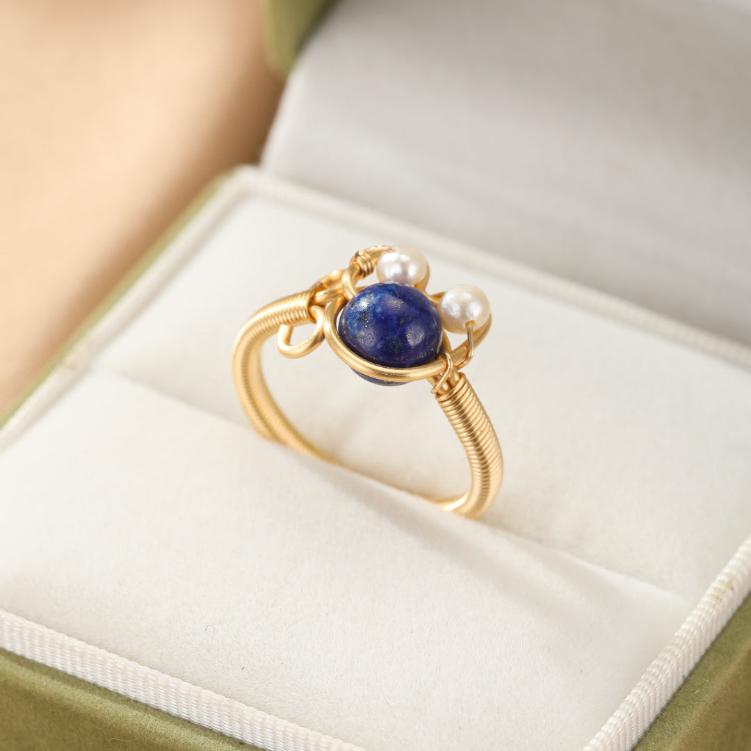Bague en fil d'or jaune sertie d'une pierre bleue lapis-lazuli et de deux petites perles blanches, présentée dans un écrin.