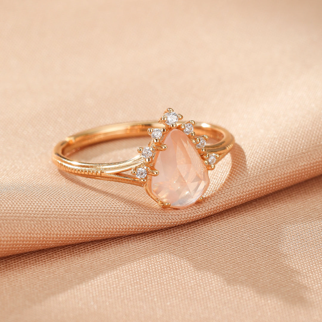 Bague en plaqué or 9K avec une pierre de quartz rose en forme de goutte, ornée de petites pierres scintillantes disposées en arche, posée sur un tissu satiné de couleur pêche.
