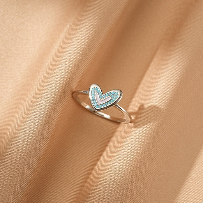 Bague en argent avec un pendentif en forme de cœur orné de petites pierres bleues et d'un cœur rose pâle au centre, posée sur un tissu satiné.