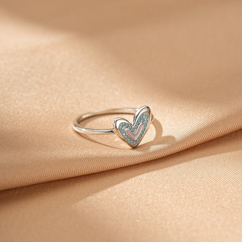 Bague en argent avec un cœur orné de petites pierres bleues et d'une bande rose au centre, posée sur un tissu satiné beige.