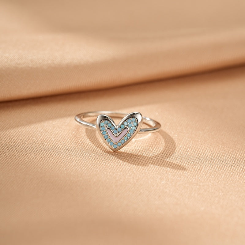 Bague cœur argent avec un cœur rose pâle incrusté de petites pierres bleues turquoise, sur un fond satiné beige.