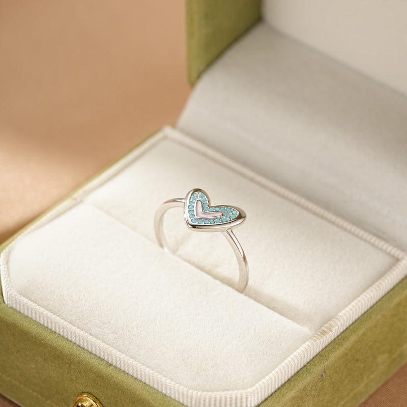 Bague en argent avec un cœur serti de petites pierres turquoise bleues et un cœur rose pâle à l'intérieur, posée dans une boîte à bague verte.