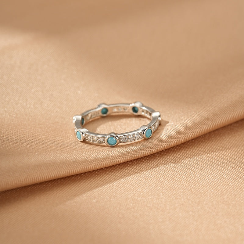 Bague fine en argent 925 ornée de petites pierres de turquoise bleues et de cristaux blancs, posée sur un tissu satiné beige.
