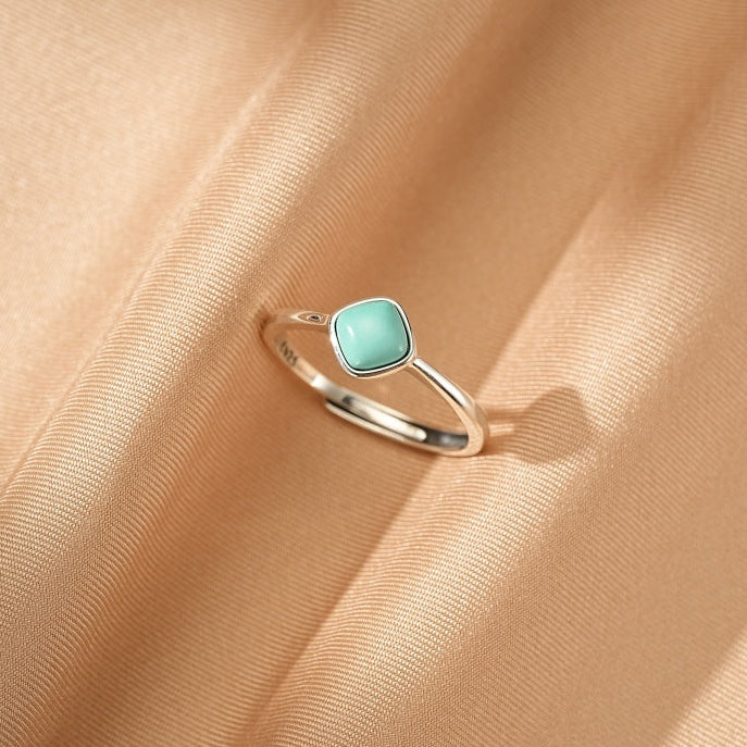 Bague minimaliste en argent 925 avec une petite pierre turquoise carrée de couleur bleu clair, posée sur un tissu satiné couleur champagne.