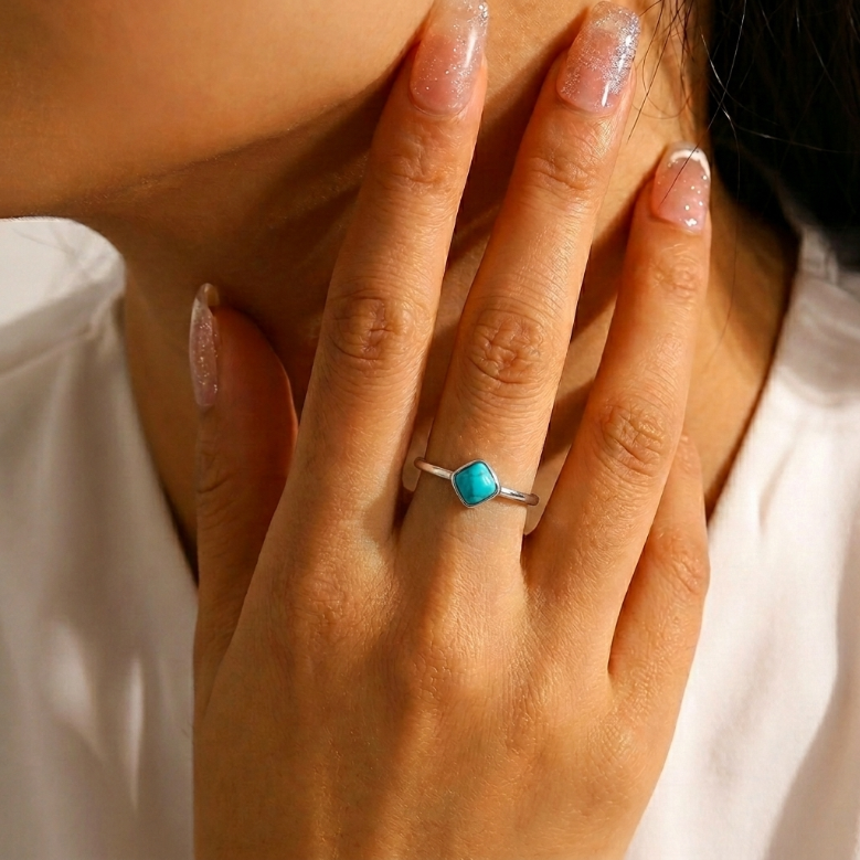 Bague fine en argent avec une turquoise carrée bleue au centre, portée à l'annulaire d'une main aux ongles manucurés.