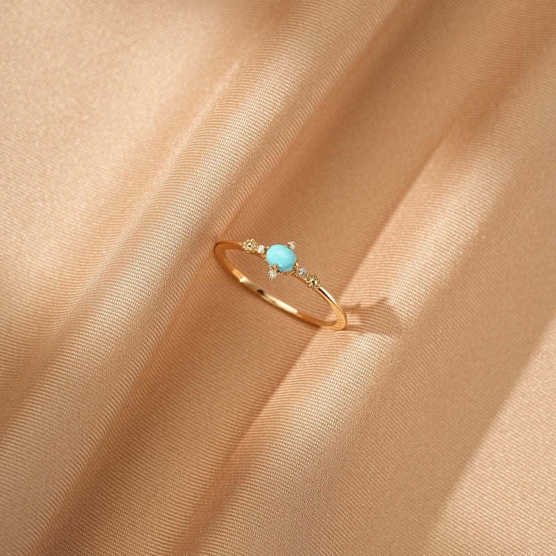 Bague fine en argent plaqué or 14k avec une turquoise centrale et de petits diamants de chaque côté, posée sur un tissu satiné beige.