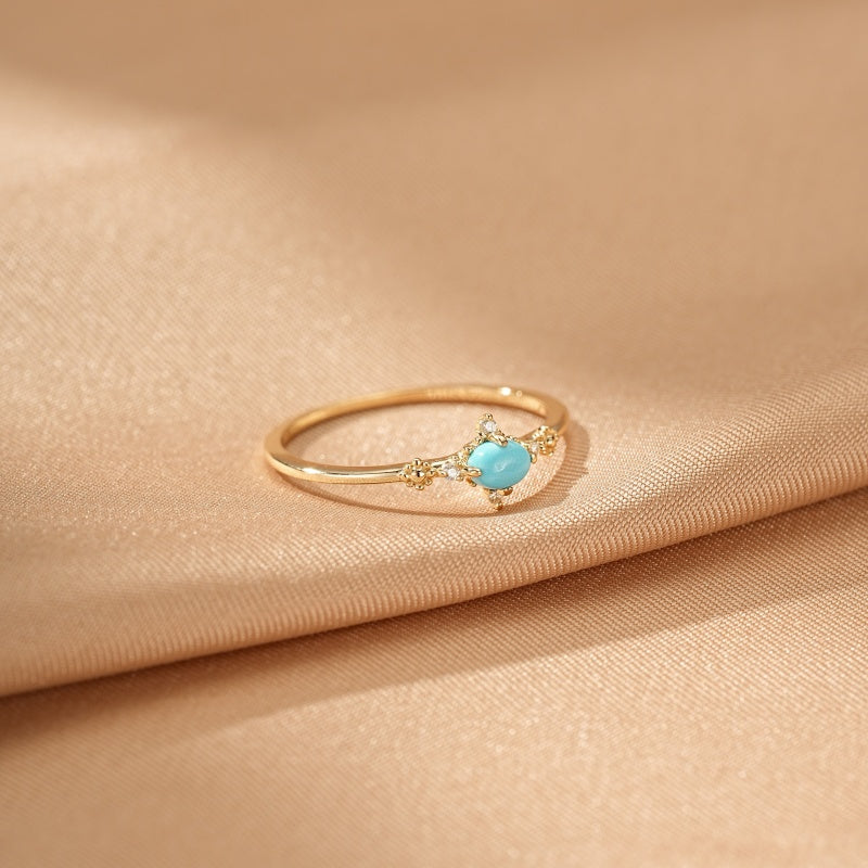 Bague en argent plaqué or 14k avec une turquoise centrale et de petites pierres scintillantes de chaque côté, posée sur un tissu soyeux beige.