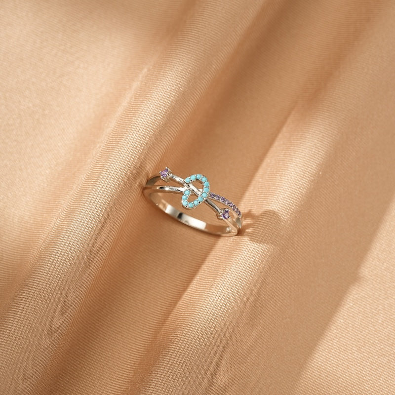 Bague fine en argent avec un motif de cœur noué serti de petites pierres turquoise et parsemé de petites pierres violettes sur un fond satiné beige