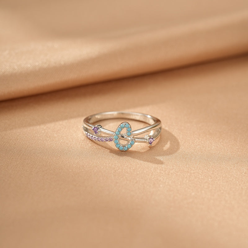Bague double anneau en argent ornée de la lettre "C" sertie de turquoise et de petites pierres violettes sur fond satiné beige.