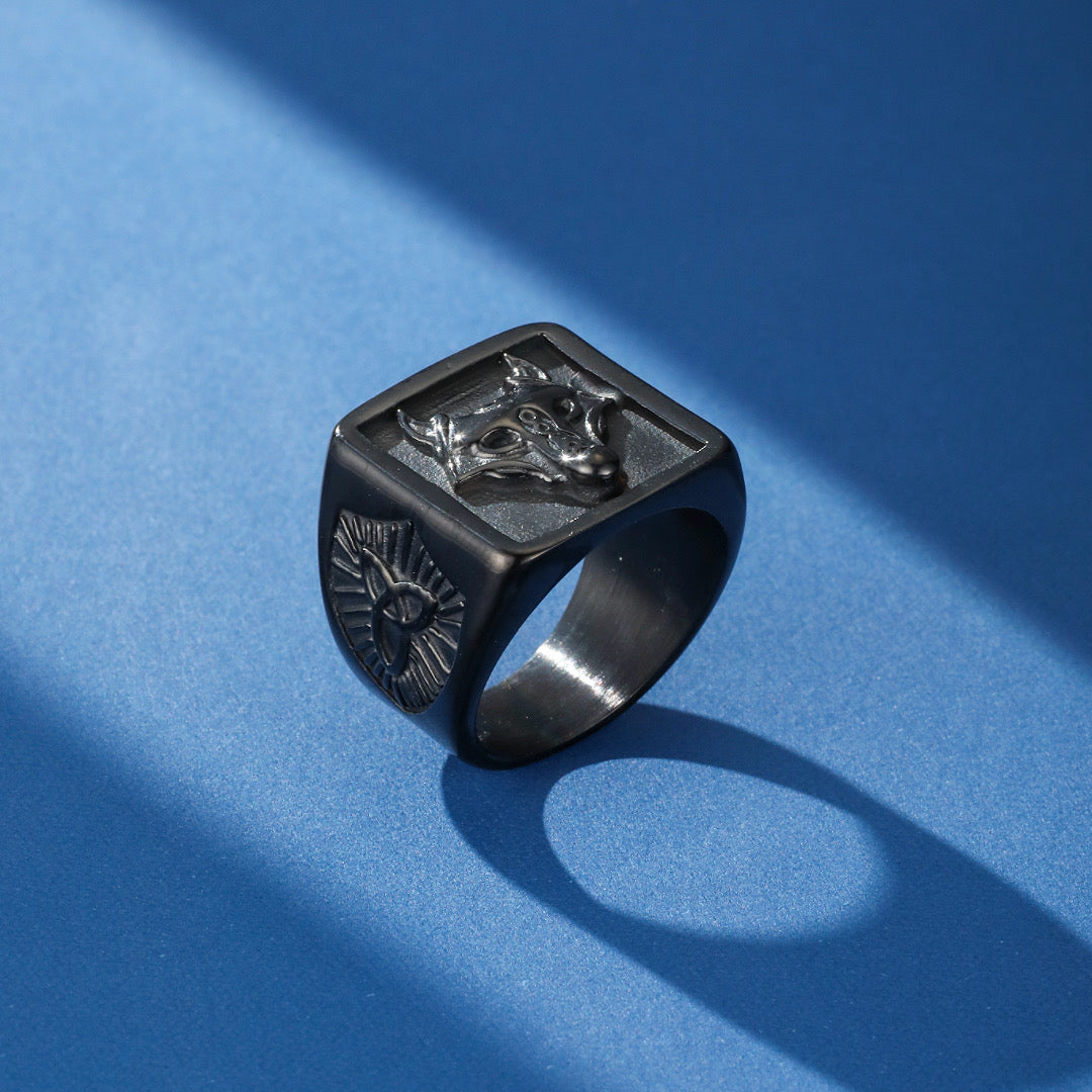 Bague viking noire en acier inoxydable avec une tête de loup gravée en relief sur le dessus et un symbole nordique sur le côté, sur un fond bleu avec des ombres.