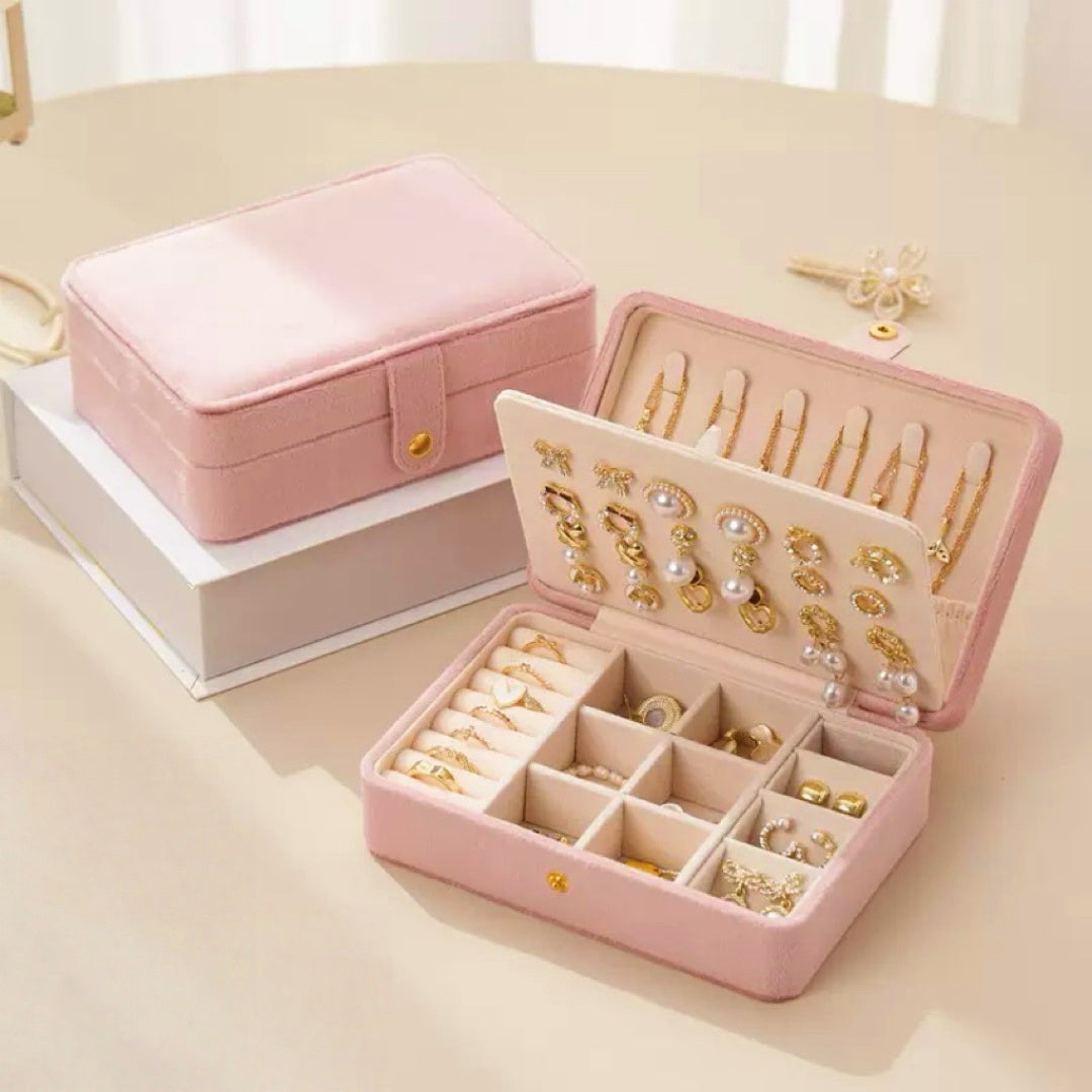 Boîte à bijoux rose en velours ouverte, révélant des compartiments remplis de colliers, bagues, boucles d'oreilles et autres accessoires de mode. Une autre boîte fermée est visible en arrière-plan.