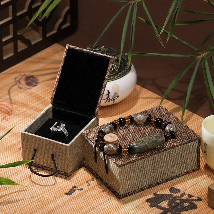 Boîte cadeau zen en lin beige ouverte avec une bague ornée de pierres précieuses, à côté d'une boîte fermée ornée d'un bracelet bouddhiste.