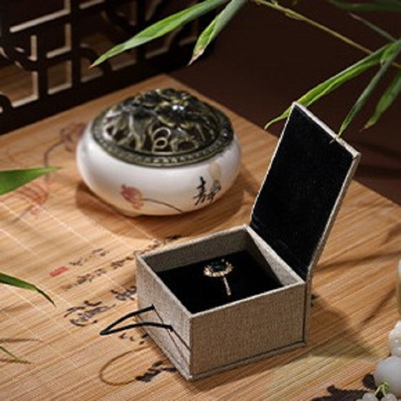 Boîte cadeau zen en lin beige, ouverte, révélant une bague avec une pierre verte au centre, posée sur un support en velours noir.