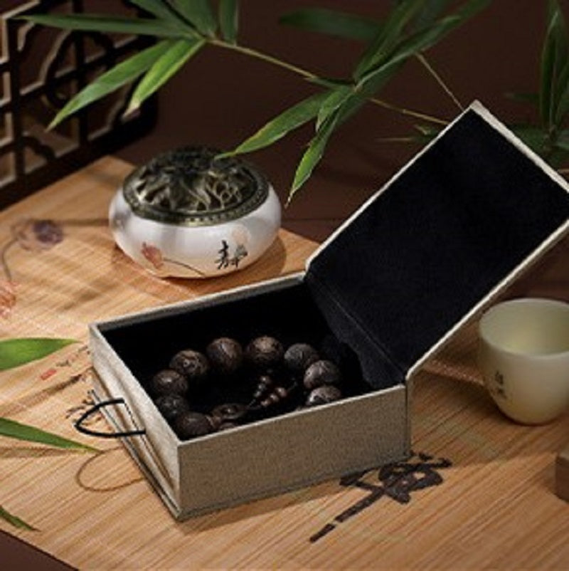 Boîte cadeau zen en lin beige, ouverte, avec un bracelet en perles sombres à l'intérieur, à côté d'un brûle-parfum et d'une tasse en céramique.