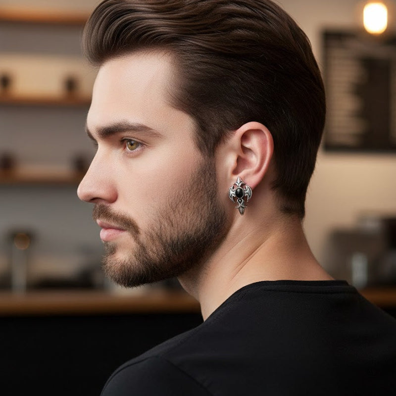 Boucle d'oreille homme argentée avec une pierre noire centrale et des motifs complexes, portée par un homme barbu.
