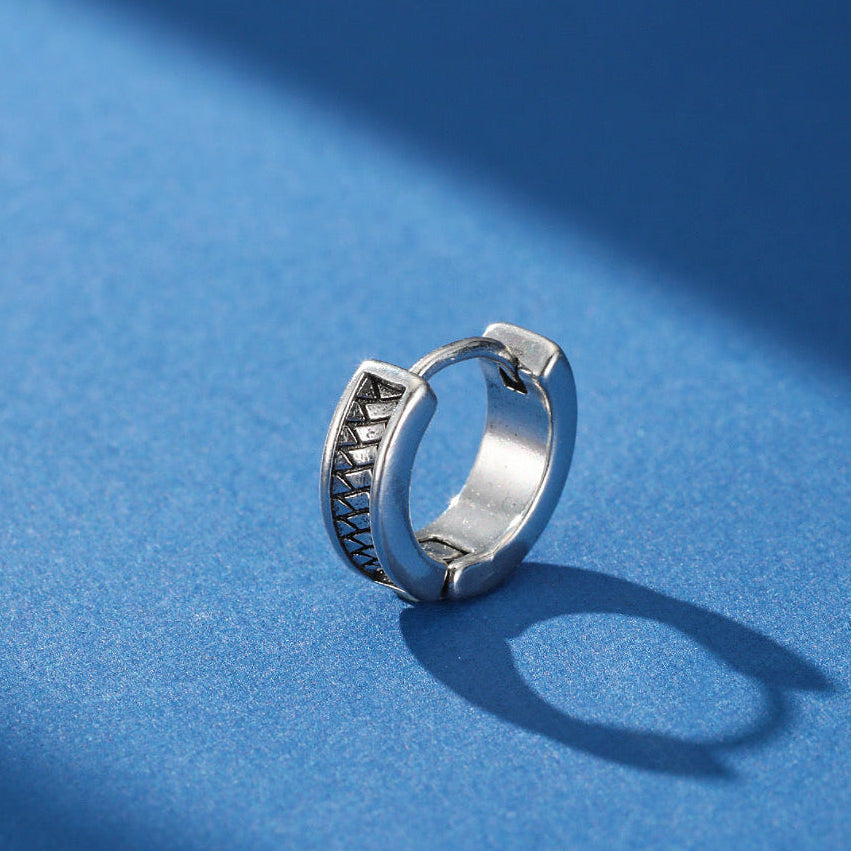 Boucle d'oreille homme argentée avec un motif géométrique rappelant une trame urbaine, sur fond bleu.