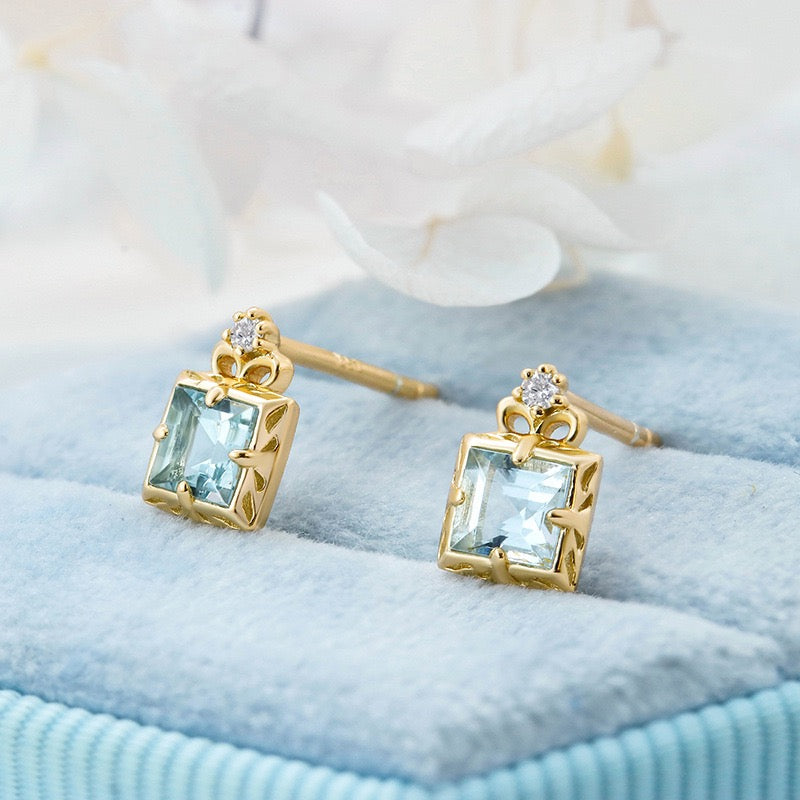 Paire de boucles d'oreilles plaqué or ornées d'une aigue-marine carrée et d'un petit diamant, posées sur un écrin bleu.
