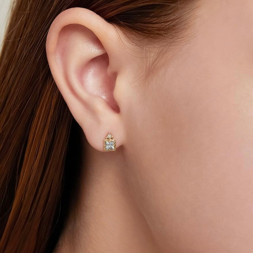 Boucle d'oreille plaqué or avec une aigue-marine carrée au centre et de petits zircons sur les côtés, portée à l'oreille.