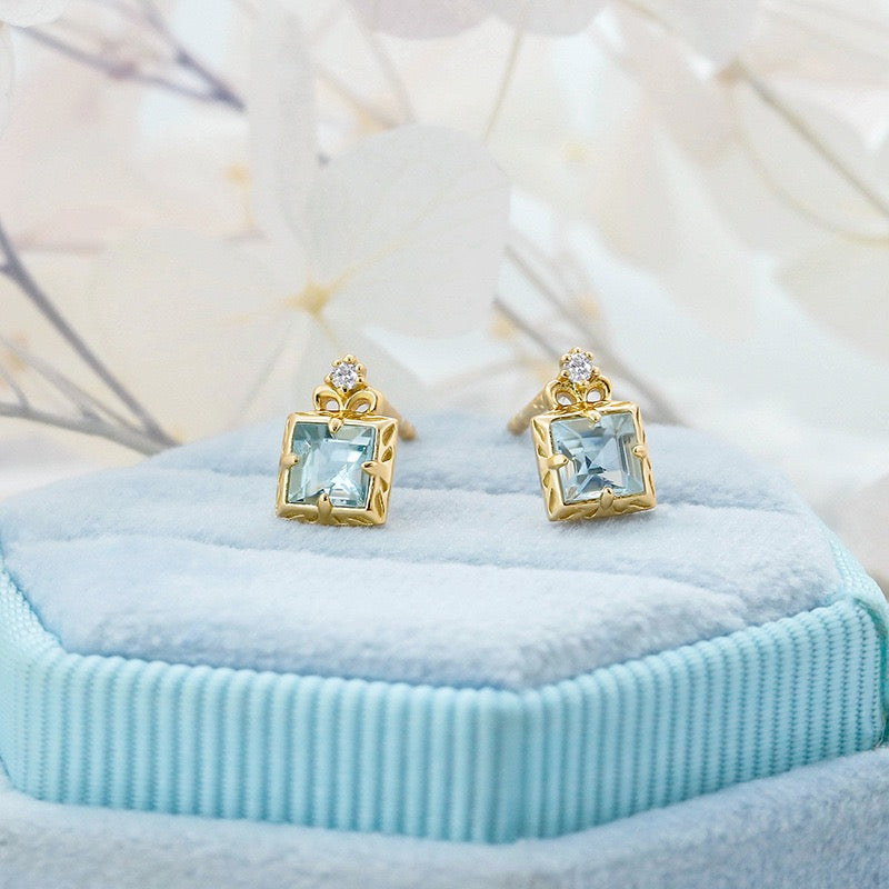 Boucles d'oreilles carrées en aigue-marine et or jaune avec un petit diamant, posées sur un écrin bleu.