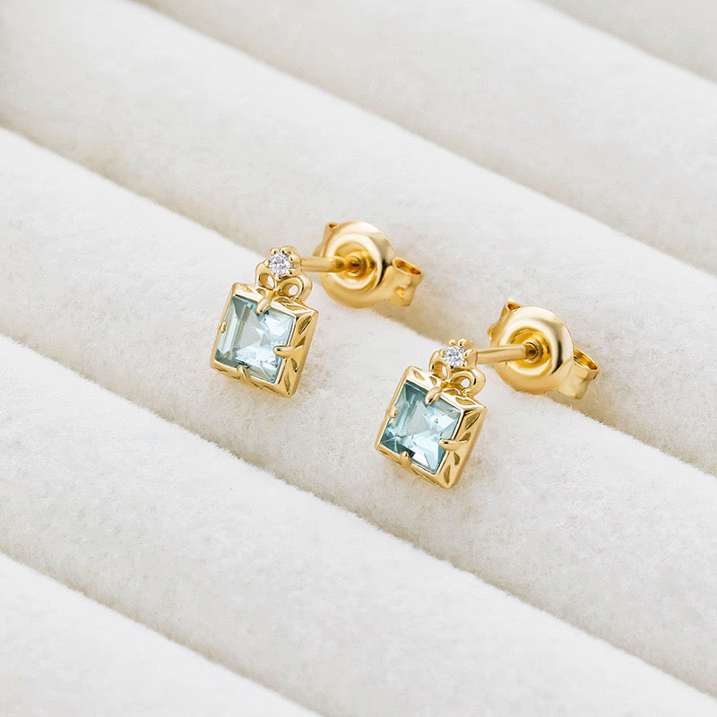 Paire de boucles d'oreilles en plaqué or ornées d'aigue-marine carrée et d'un petit diamant, sur fond blanc texturé.