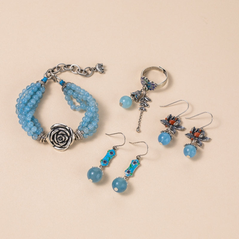 Ensemble de bijoux en aigue-marine composé d'un bracelet à plusieurs rangs orné d'une rose en argent, de deux paires de boucles d'oreilles pendantes avec des perles bleues et des motifs floraux bleus et rouges, et d'une bague ornée d'une perle bleue et de papillons.