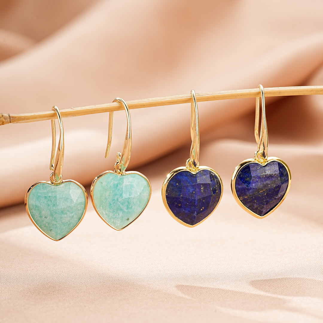 Deux paires de boucles d'oreilles en forme de cœur, l'une en amazonite bleu-vert pâle, l'autre en lapis-lazuli bleu profond, suspendues à des crochets dorés sur un fond rose pâle.