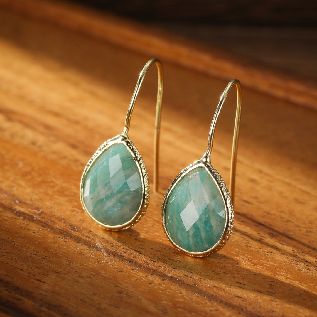 Boucles d'oreilles pendantes en amazonite verte sculptée en forme de goutte d'eau, serties dans un cadre doré texturé, sur un fond en bois naturel.