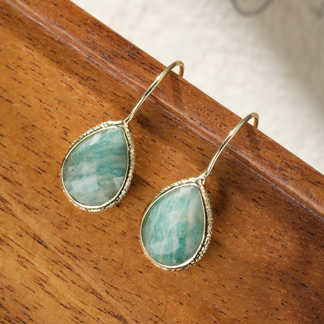 Boucles d'oreilles pendantes en amazonite verte, en forme de goutte, serties dans un cadre doré texturé, sur un fond en bois.