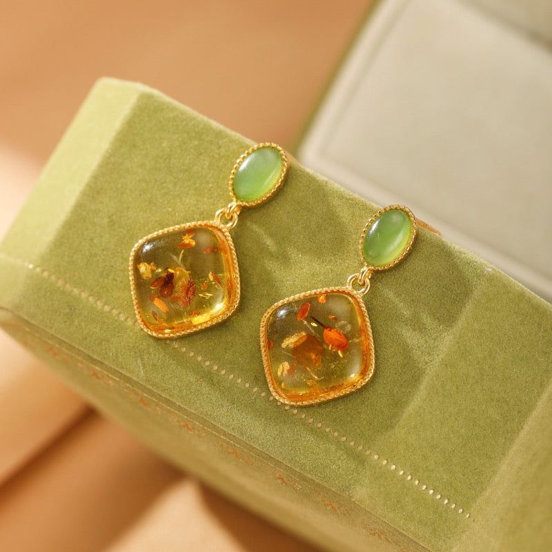 Boucles d'oreilles pendantes Feu Doré composées d'une pierre de jade verte ovale et d'une pierre d'ambre carrée en argent plaqué or, posées sur un écrin vert.