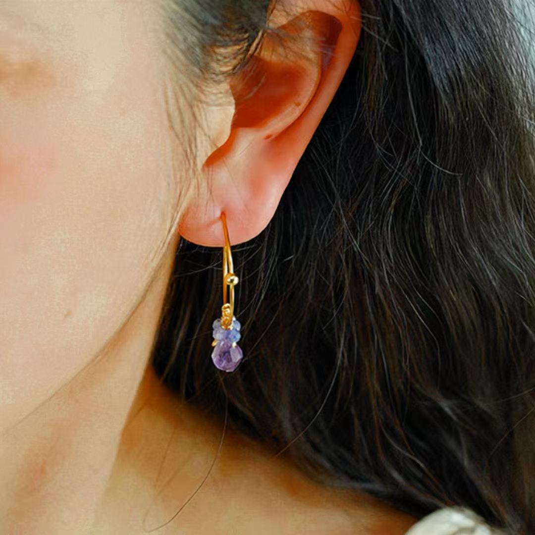 Gros plan sur une boucle d'oreille pendante en argent plaqué or, ornée de deux pierres facettées, une améthyste violette et une tanzanite bleue, portée à l'oreille d'une femme.