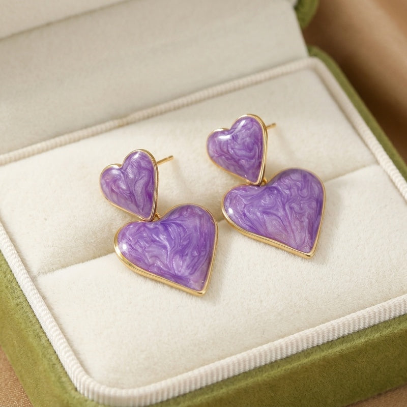 Boucles d'oreilles cœur violettes avec un motif marbré, montées sur un support doré, présentées dans un écrin vert.
