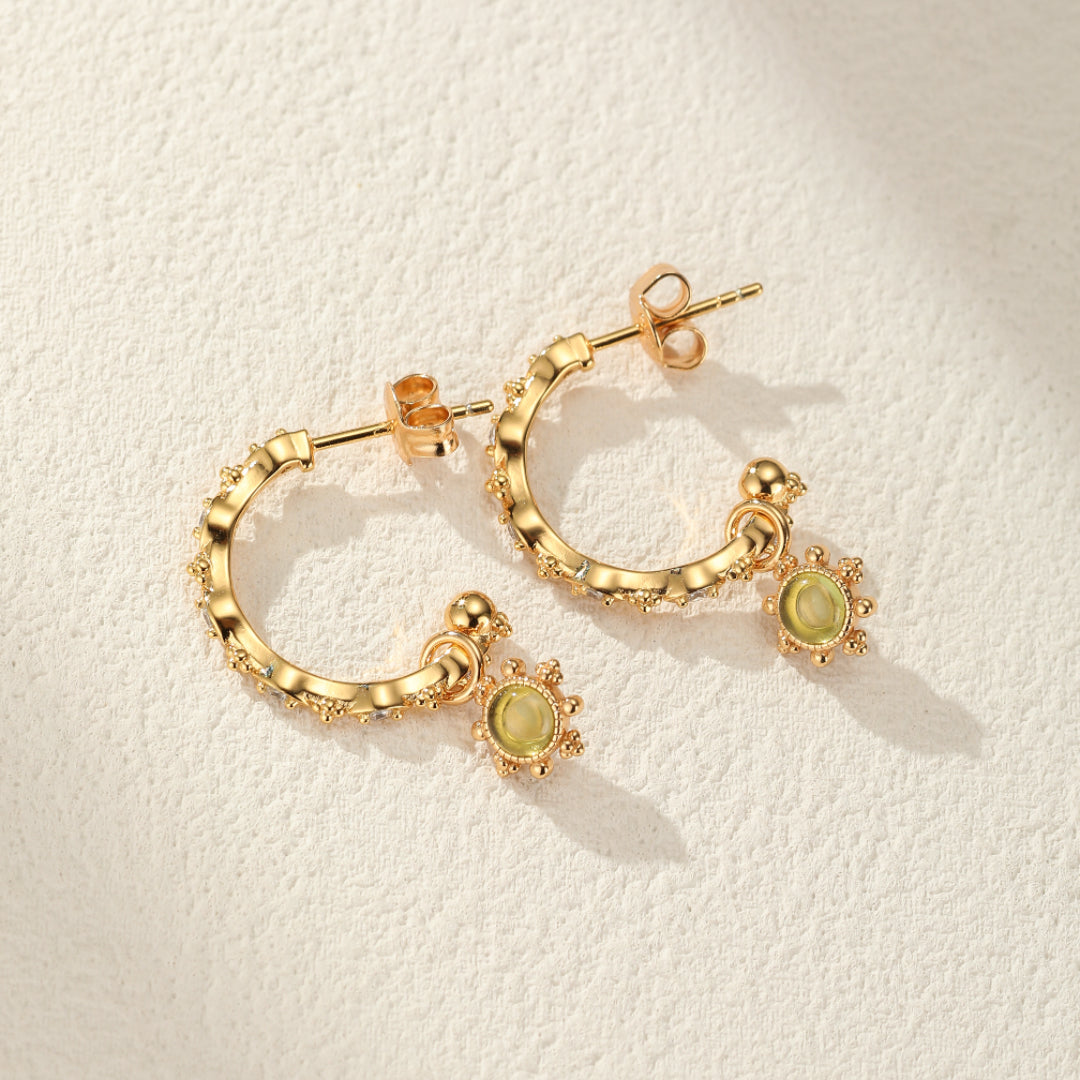 Boucles d’oreilles créoles plaquées or avec des pierres de péridot rondes et un motif soleil, sur fond texturé clair.