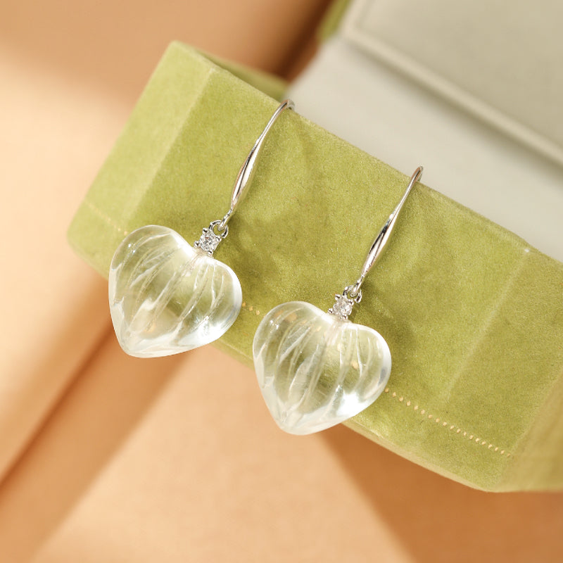 Boucles d'oreilles en forme de cœur en cristal de roche transparent, suspendues à des fils argentés ornés d'une petite pierre scintillante, présentées dans un écrin vert.