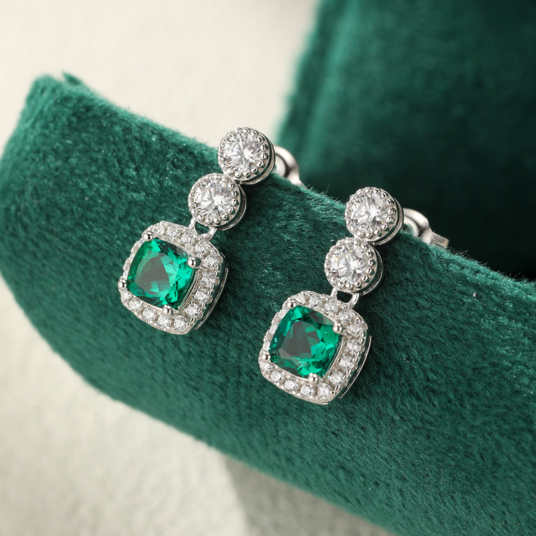 Boucles d’oreilles pendantes en argent plaqué platine avec des émeraudes carrées serties d’éclatants cristaux blancs sur un fond vert velours vert.