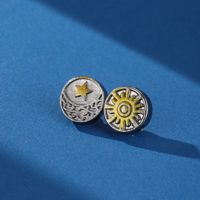 Paire de boucles d'oreilles homme Duo Cosmique avec un motif lune et étoile sur une boucle et un soleil stylisé sur l'autre, sur fond bleu.