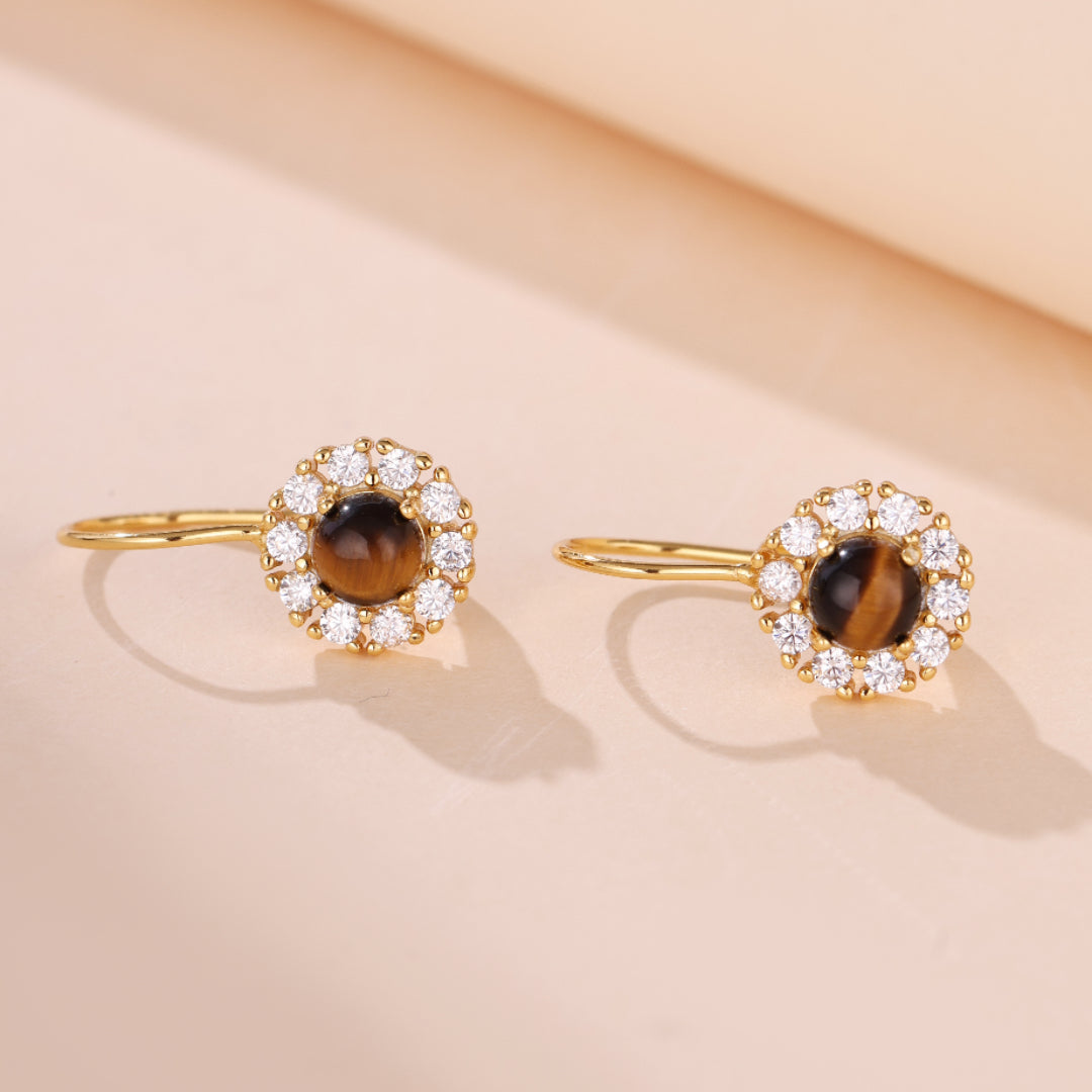 Paire de boucles d'oreilles en argent plaqué or ornées d'un œil de tigre rond entouré de petits cristaux brillants, sur fond clair.