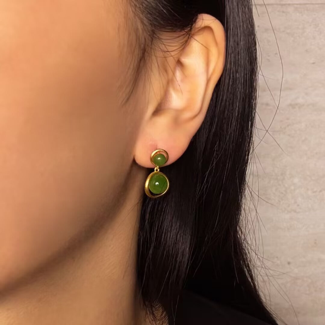 Boucles d’oreilles pendantes en argent plaqué or avec deux pierres de jade rondes de couleur verte, portées par une femme.