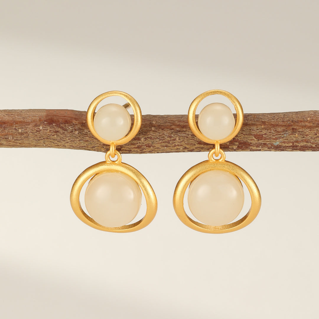 Paire de boucles d'oreilles pendantes en argent plaqué or avec des perles rondes blanches, suspendues à un support en bois.