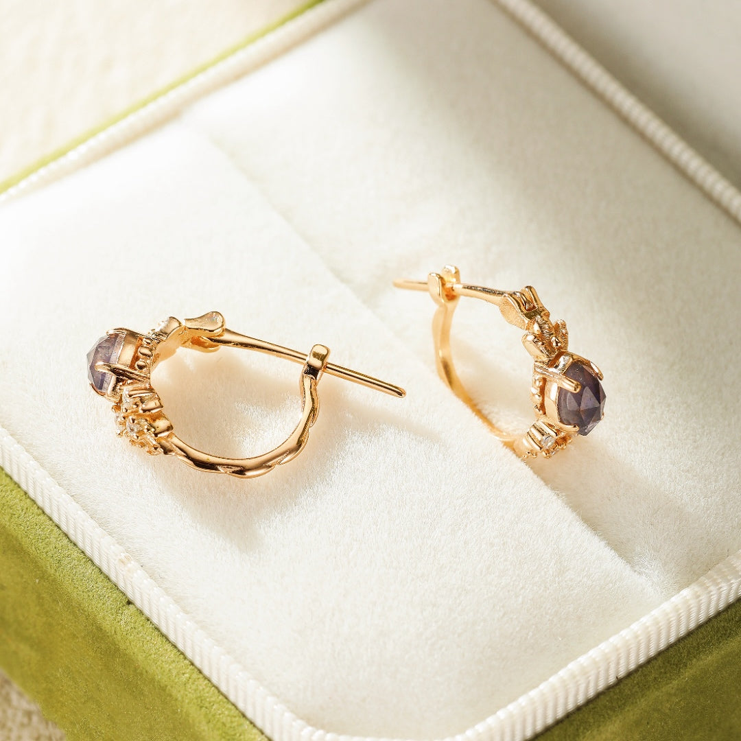 Boucles d'oreilles créoles plaquées or avec une pierre centrale violette de cordiérite et de petites pierres scintillantes, posées dans un écrin vert.