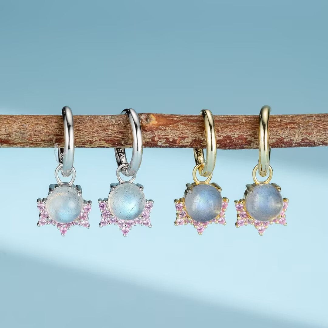 Deux paires de boucles d'oreilles pendantes en argent plaqué or ornées de pierres de lune bleues et entourées de petites pierres roses, suspendues à une branche.