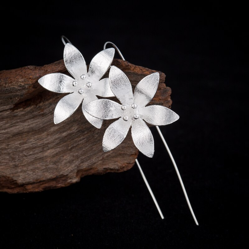 Boucles d'oreilles pendantes en argent représentant deux fleurs de lotus écloses avec des détails de pétales texturés et de petites perles au centre, posées sur un morceau de bois brut sur fond noir.